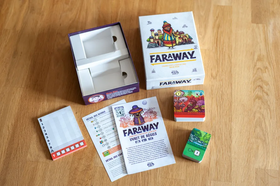 Caja de Faraway abierta junto al mazo de cartas de región, el de santuarios, la libreta de puntuación y el reglamento sobre una mesa de madera.
