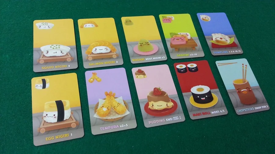 Cartas de Sushi Go! extendidas sobre una mesa verde: nigiri de calamar, de salmón y de huevo, wasabi, sashimi, tempura, pudin, rollos de maki y palillos.