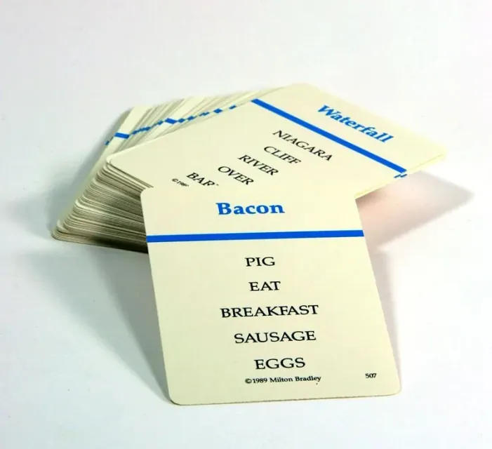 Dos cartas de Taboo con la palabra grande arriba y cinco palabras prohibidas debajo, sobre una pila de cartas.